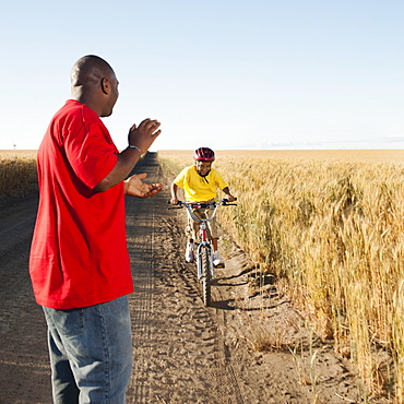 Father clapping as son (8-9) is cycling along dirt road in fields