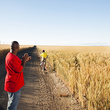 Father clapping as son (8-9) is cycling along dirt road in fields