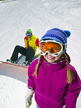 Daughter (10-11) posing with father sitting on ground with snowboard in background 