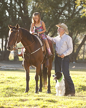 Senior man with dog assisting granddaughter (8-9) horseback riding in ranch