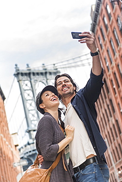 Couple taking selfie on street, Brooklyn Bridge in background, Brooklyn, New York