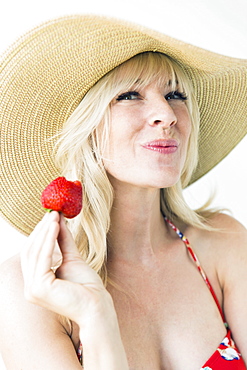 Studio portrait of blonde woman with sun hat holding strawberry