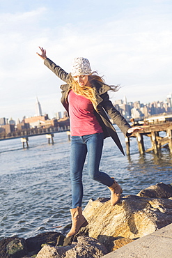 Blond woman stepping on stones by river, skyline in background, USA, New York City, Brooklyn, Williamsburg