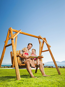 Father with daughters (2-3, 4-5) on swing reading book, USA, Utah, Garden City