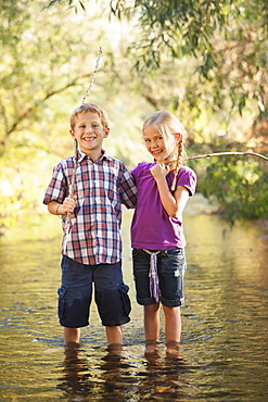 Little boy (6-7) and little girl (4-5) standing together in small steam holding wooden stick fishing poles, Lehi, Utah