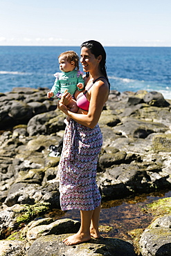 Woman holding baby daughter on rocks at beach