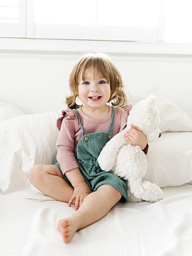 Girl (2-3) sitting on bed with teddy bear