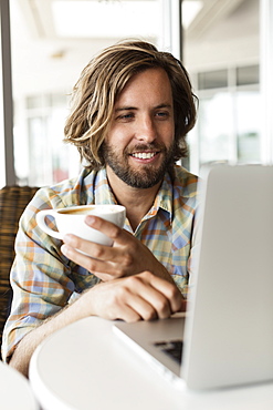 Mid adult man in coffee shop, drinking coffee, using laptop