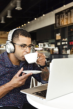 Young man in coffee shop, drinking coffee, using laptop