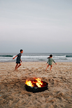 Boy (8-9) and girl (2-3) playing on beach near bonfire