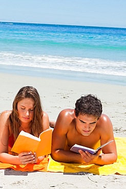 Tanned couple reading a book while lying on the beach