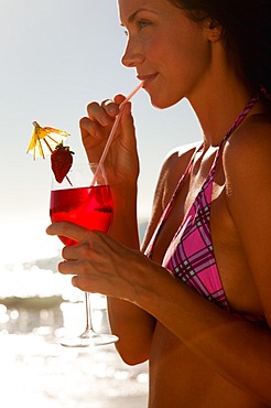 Woman using a straw to drink a cocktail while she stands on a beach