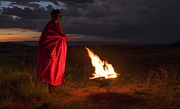 Masai Mara man wearing traditional tribal red blanket by camp fire, Masai Mara National Park, Kenya, East Africa, Africa