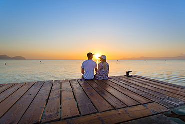 Young couple sitting at the end of a jetty in the morning atmosphere and enjoying the view of the sunrise. Playa de Muro beach, Alcudia, Mallorca, Balearic Islands, Spain