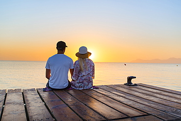 Young couple sitting at the end of a jetty in the morning atmosphere and enjoying the view of the sunrise. Playa de Muro beach, Alcudia, Mallorca, Balearic Islands, Spain