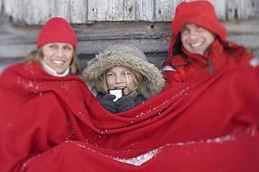 Parents and son leaning on wooden hut, warming with red blanket