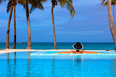 A woman in a black, white-rimmed hat, her back to the camera, rests on the side of a pool with an ocean view. Antigua, West Indies.