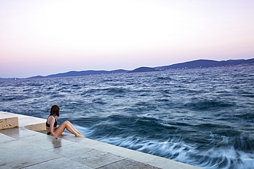 Young woman in bikini sitting on steps of the sea promenade at sunset, Zadar, Zadar, Croatia, Europe