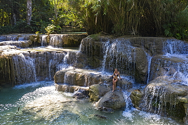 Aerial view of a beautiful young blonde woman on a rock in the natural pools of the magnificent Kuang Si Falls, Kuang Si, Luang Prabang Province, Laos, Asia