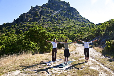 Three persons doing yoga in front of a mountain in the light of the morning sun, Jerzu, Sardinia, Italy, Europe