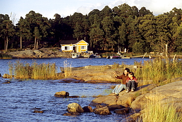 A man and a child sitting at the shore of a skerry in the light of the evening sun, South Finland, Europe