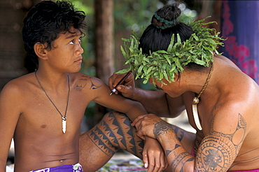 A man wearing a headdress tatooing a boy at Tiki village, Moorea, French Polynesia, Oceania