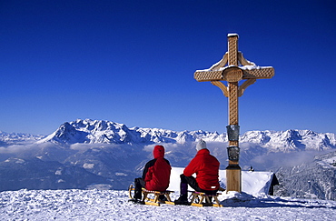 Two persons sledging at cross of Bischlinghoehe, view to Hochkoenig range, skiing area Werfenweng, Salzburg, Austria