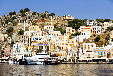 Yacht and sailing boats anchoring at quay of harbour Gialos, picturesque mansions at mountainside in background, Simi, Symi Island, Greece