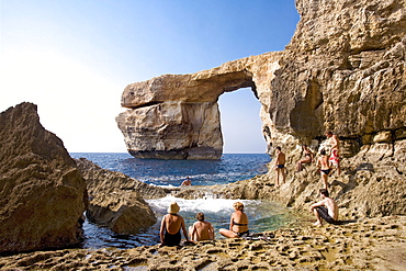 People sitting on rocks at the sea, rock arch in the background, Gozo, Malta, Europe