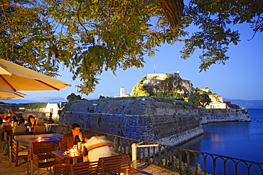 People sitting in the cafe Akteion in the evening, in the background the old citadel, Corfu, Ionian Islands, Greece