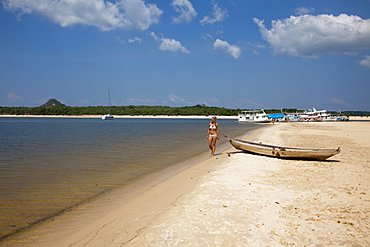 Woman strolling along the beach on a side arm of the Amazon river with Amazon river boats in the distance, Alter do Chao, Para, Brazil