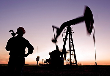 Worker holding a pipe wrench looking out over a moving oil pumpjack, near Carlyle, Saskatchewan