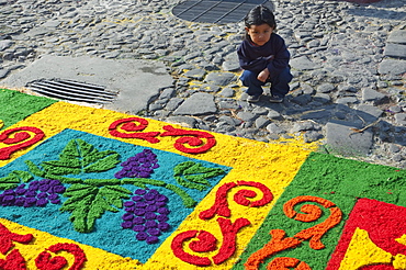 Girl by a carpet made of sand & sawdust along the Good Friday processional route. Carpet-making is thought of as a sacrificial act, as the elaborate detail and time that go into the carpet making is a way for people to give something of themselves in memory of the crucifixion of Jesus. These carpets last on average 2 hours before they are destroyed by the many feet that march over them during a procession in Antigua Guatemala., Sacatepuquez, Guatemala