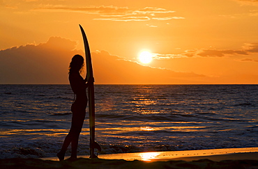 Hawaii, Female surfer on beach silhouetted against orange sunset over ocean.