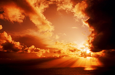 Orange sunset over ocean, Sunrays bursting through puffy clouds.