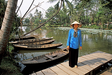 Vietnam, Mekong Delta, Woman in traditional clothing, standing on dock near river and old boats.