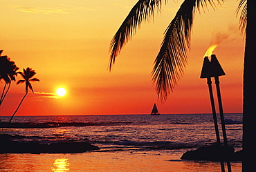 Hawaii, Big Island, Kohala, Waiulua Bay, Orange sunset with palm trees, sailboat and tiki torches.