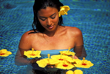 Young Polynesian woman in pool surrounded by floating yellow hibiscus looking down