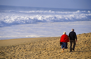 View from behind senior couple walking on beach, arm in arm white shorebreak ocean water