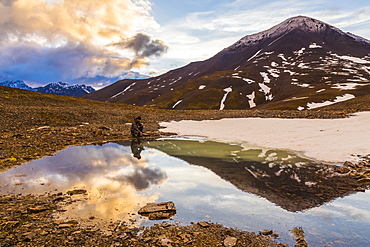 A backpacker pauses for rest beside a puddle while crossing over an unnamed pass in the Brooks Range under the midnight sun at 12:14 a.m.; Alaska, United States of America