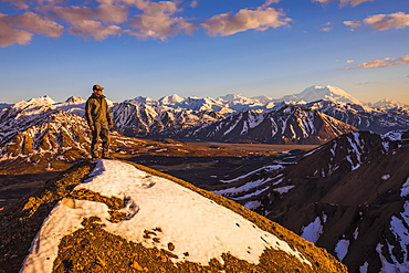 A man on a snowy ridge near Polychrome Mountain in Denali National Park faces the sunset with Denali in the background; Alaska, United States of America