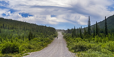 Two cyclists ride down a gravel road between hills and forest; Canada