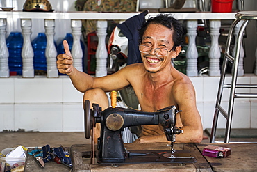 Tailor, shirtless and sitting by his sewing machine giving the thumbs up with a smile, My Long, Mekong Delta, Tien Giang, Vietnam