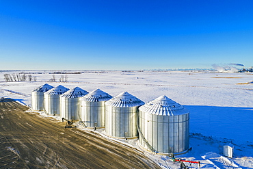 Aerial view of snow-covered large metal grain storage bins in a snow-covered field with blue sky, South of Calgary, Alberta, Canada