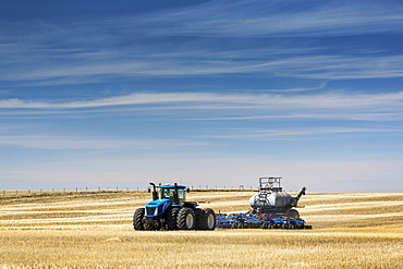 Tractor with air seeder, seeding a stubble field with blue sky and hazy clouds, near Beiseker, Alberta, Canada