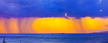 Storm clouds with rain and clouds glowing a bright yellow at sunset off Waikiki Beach with a silhouetted sailboats in the water, Honolulu, Oahu, Hawaii, United States of America