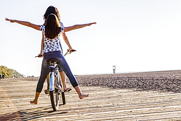 Two friends riding tandem on a bike together with bare feet down a wooden boardwalk along Woodbine Beach, Toronto, Ontario, Canada