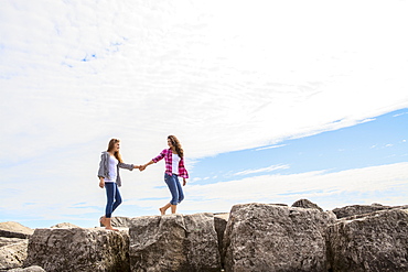 Two teenage girls holding hands as they walk along boulders, Woodbine Beach, Toronto, Ontario, Canada