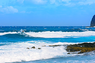 A surfer on the breaking waves close to the shore of Kaupo Cove, with bright blue water and sky on the horizon, Oahu, Hawaii, United States of America