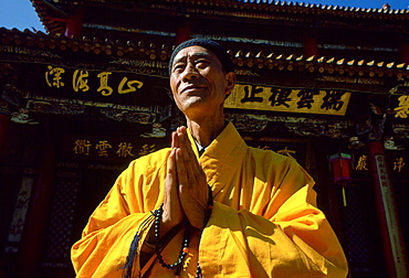 A Buddhist monk in saffron coloured robes with hands together while praying at the Buddhist Temple in Huating, China